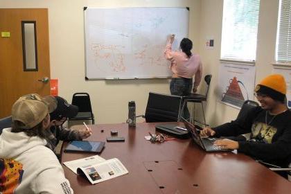 students sit at a table while one student writes on a white board